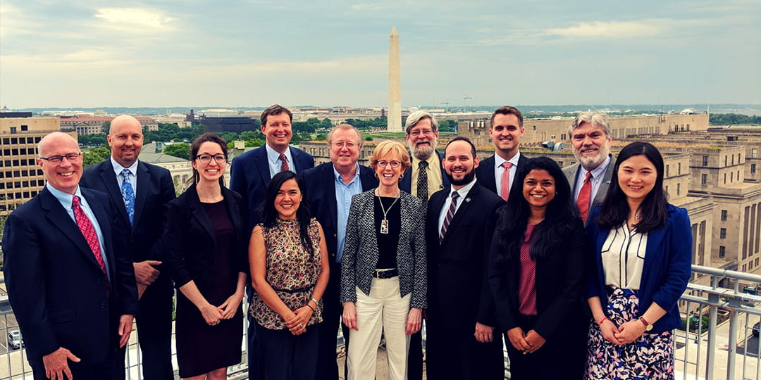 Staff and faculty of the GW Regulatory Studies Center standing on a rooftop with the Washington Monument in the background